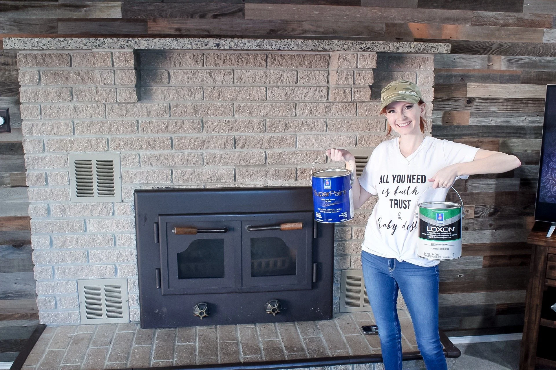 Corinne of Slayathomemother.com holding up paint cans in front of my tan fireplace, ready to paint!